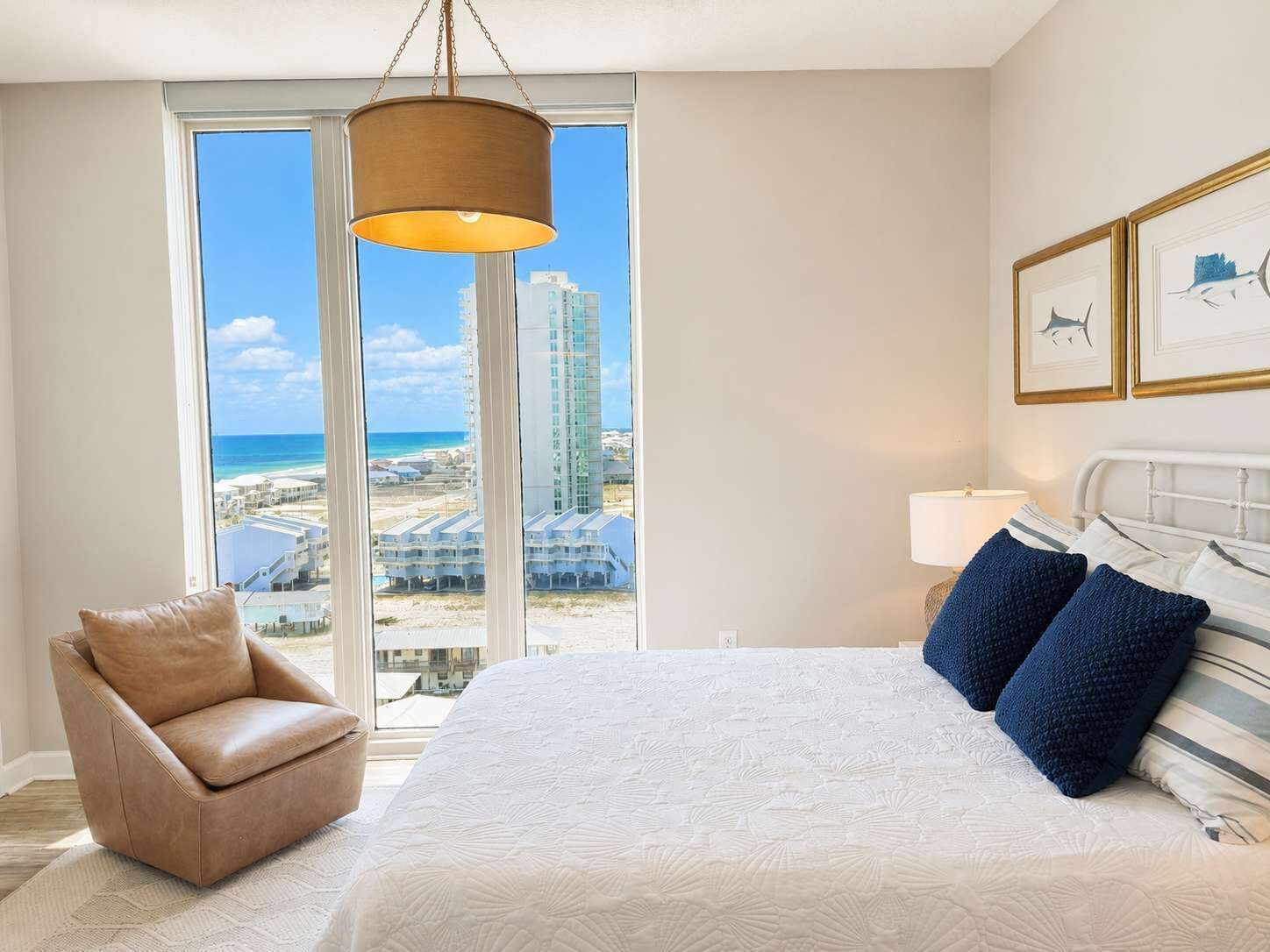 A bedroom with a large bed, a chair, a hanging light fixture, and a view of a beach and buildings.