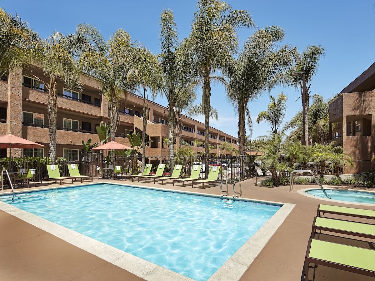 A pool area with multiple lounge chairs and umbrellas, surrounded by palm trees and a multi-story building.