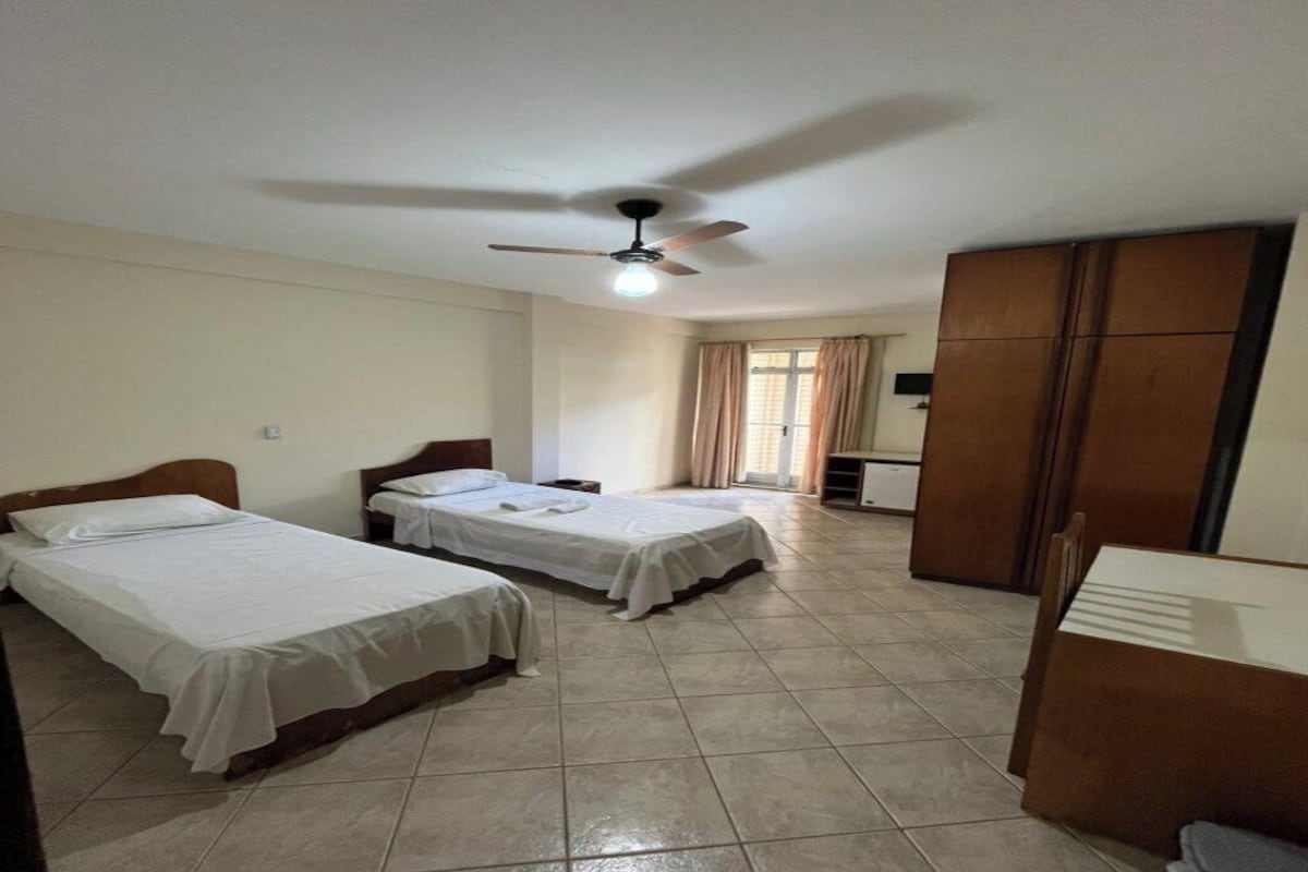 Habitación de hotel con dos camas individuales, ventilador de techo, armario y una mesita pequeña.