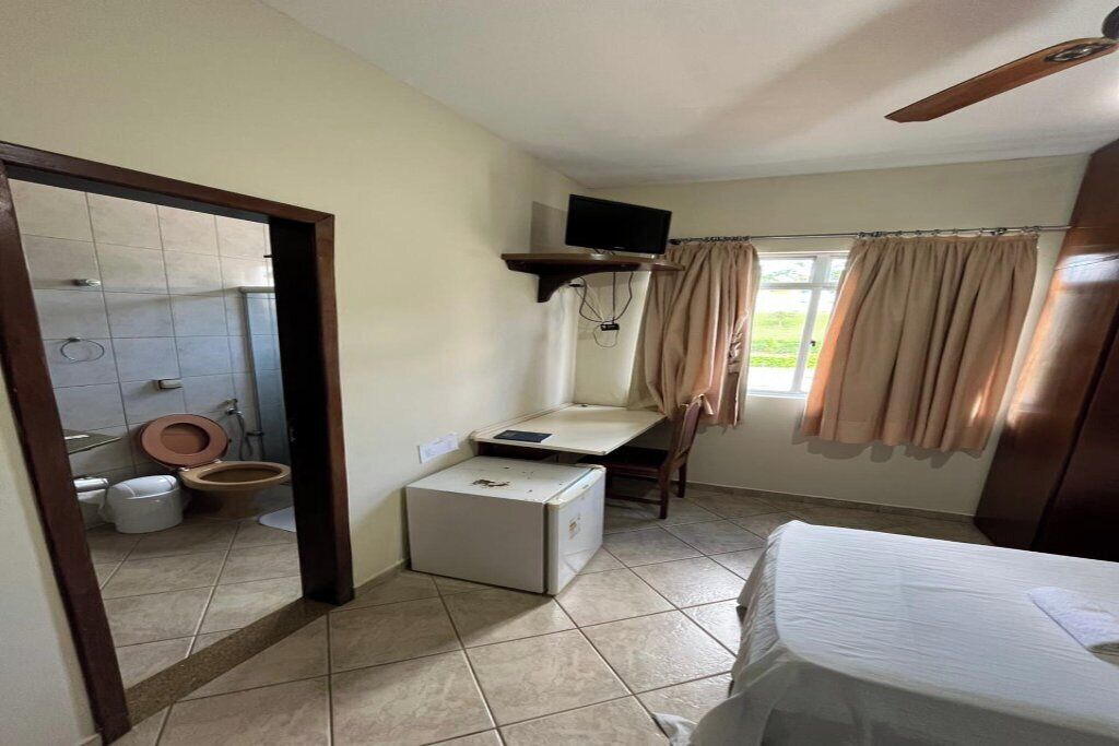 Una habitación pequeña y funcional con cama, escritorio con computadora, televisor montado en la pared y ventana con cortinas.