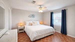 Un dormitorio con cama, mesitas de noche, ventilador de techo, ventana con cortinas y un adorno en la pared.