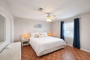 Ein Schlafzimmer mit einem Bett, Nachttischen, einem Deckenventilator, einem Fenster mit Vorhängen und einem Wandbild.