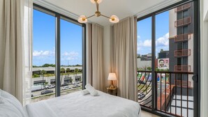 Ein Hotelzimmer mit einem großen Bett, einem Nachttisch, einer Lampe und Blick auf die Stadt durch große Fenster.