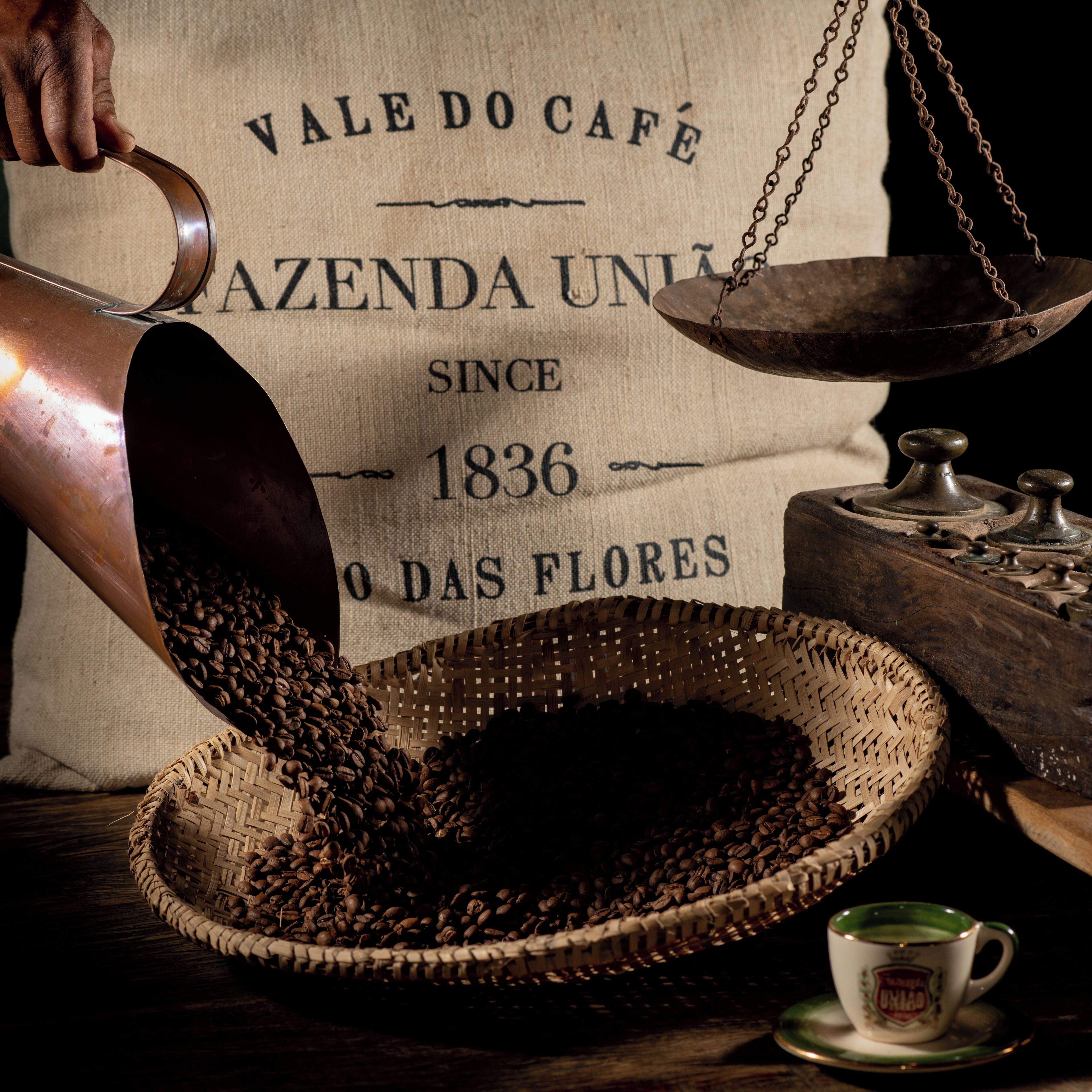 Un saco de arpillera con texto y una balanza, granos de café derramándose en una cesta tejida, y una taza sobre un platillo.