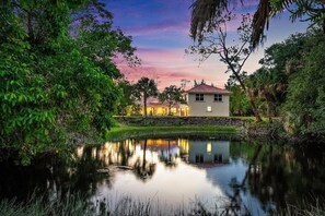 A house with a pond in front, surrounded by trees and a sunset sky.