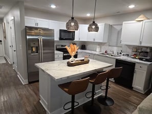 Una cocina moderna con una isla central, taburetes de bar, electrodomésticos de acero inoxidable, iluminación colgante y armarios blancos.