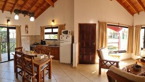 Amplia sala de estar con mesa de comedor de madera, un sofá y un televisor. Cuenta con una zona de cocina equipada con refrigerador y microondas. El ambiente se caracteriza por techos altos con vigas de madera y ventanas grandes.