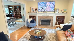A living room with a fireplace, a large TV, a dining area, and a bookshelf. - October in Boston - Jamaica Plain Home Near Hospitals & New England Adventures (Boston)