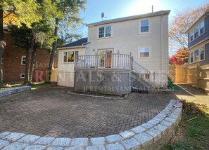 Ein zweistöckiges Haus mit gepflasterter Terrasse, einer kleinen Holzterrasse und einer Ziegelwand.