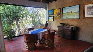 A dining area with a table set for two, surrounded by wicker chairs, a wooden cabinet, and a view of greenery outside. - Villa Oaxaca, a stately residence with 6 suites, art, culture, nature, and a pool (Oaxaca de Juárez)