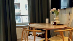 Une pièce moderne avec une table ronde en bois, quatre chaises en osier, un téléviseur à écran plat fixé au mur et une fenêtre avec des rideaux foncés.