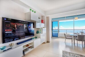 Ein modernes Wohnzimmer mit großem Flachbildfernseher, einem Essbereich mit Tisch und Stühlen und Blick zum Himmel durch große Fenster.