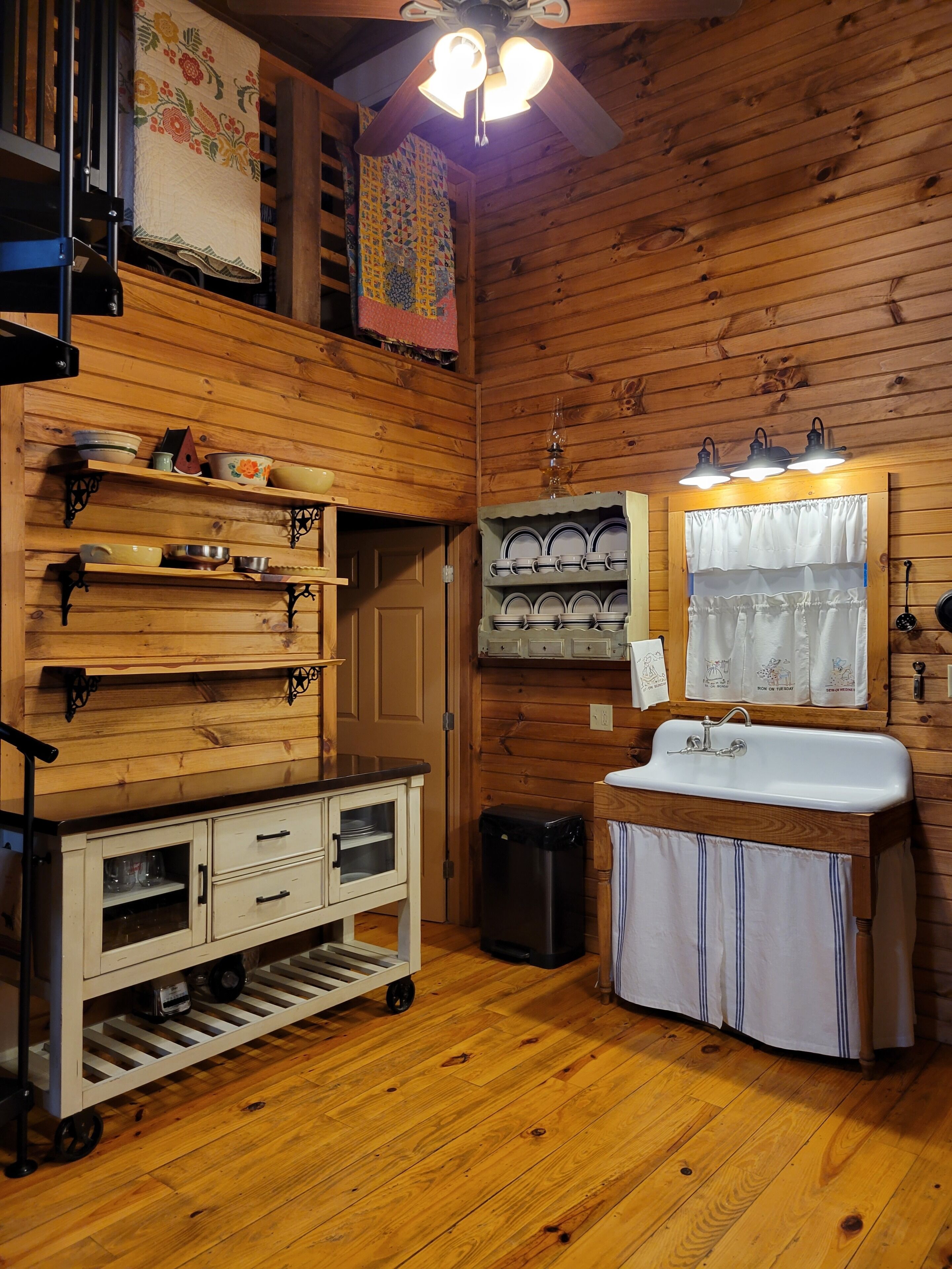 A wooden cabin interior with a kitchenette, shelves with dishes, a ceiling fan, and a window with curtains.