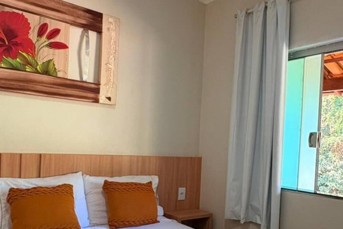 A hotel room with a bed, a painting, a window with curtains, and a view of greenery outside.