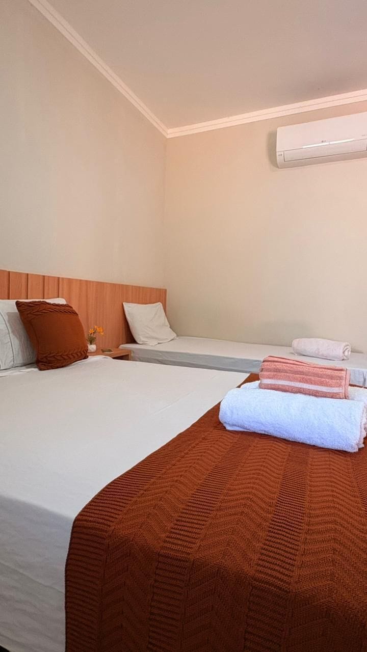 A hotel room with two beds, a wooden headboard, a wall-mounted air conditioner, and a small table with a vase of flowers.