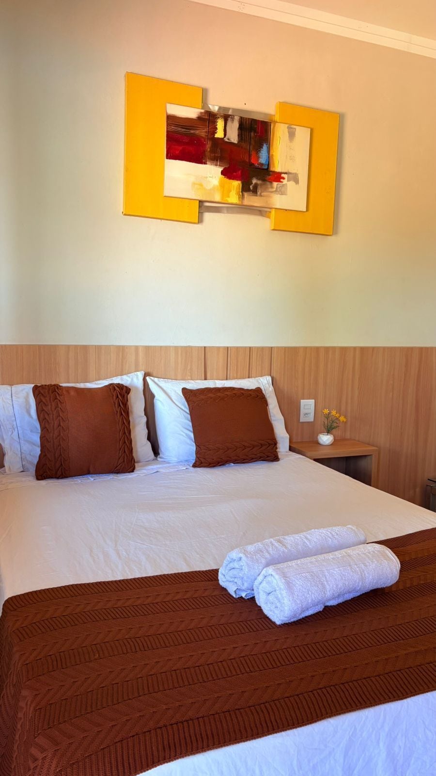 A hotel room with a large bed, brown and white bedding, a wooden headboard, a nightstand with a vase of flowers, and a painting on the wall.