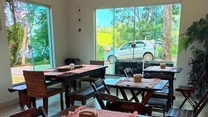 Uma área de refeições com mesas e cadeiras de madeira, grandes janelas com vista para um carro e vegetação lá fora.