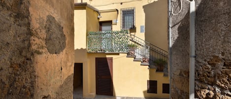 A narrow alleyway with a stone floor, a metal railing, and a building with a yellow facade and a window with a metal grille.