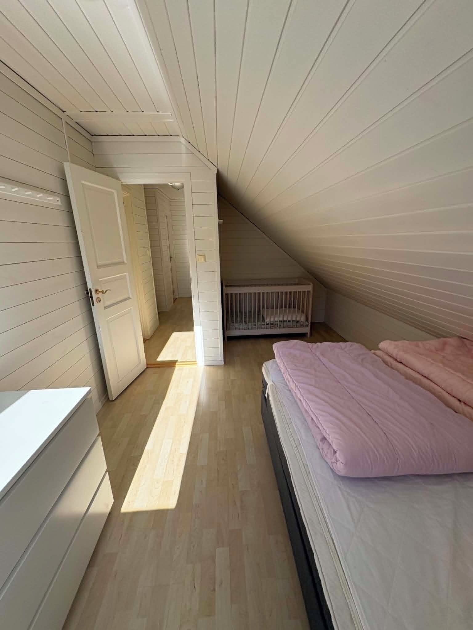 Una habitación con techo inclinado, cama con ropa de cama rosa, un radiador y una puerta que da a otra habitación.