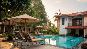 A swimming pool with lounge chairs and umbrellas, surrounded by greenery and a building with a tiled roof. - Seahorse Villas - Three-Bedroom Apartment (NEGOMBO)