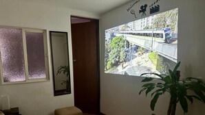 Quarto com piso de madeira, sofá bege, um pequeno banquinho, planta em vaso, TV fixada na parede e porta que dá para outro cômodo.