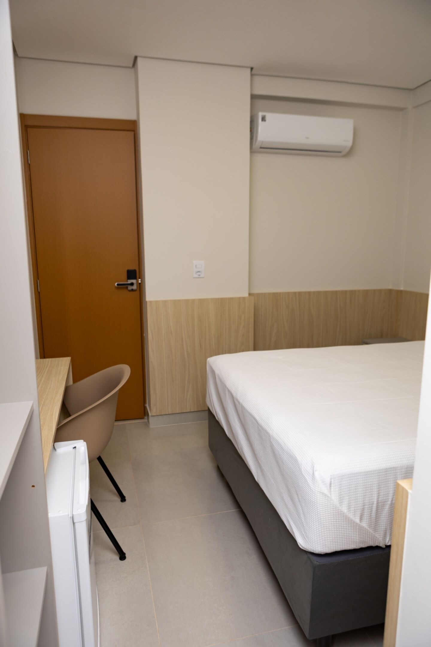 A small, neatly arranged bedroom with a bed, a chair, a desk, and an air conditioning unit.