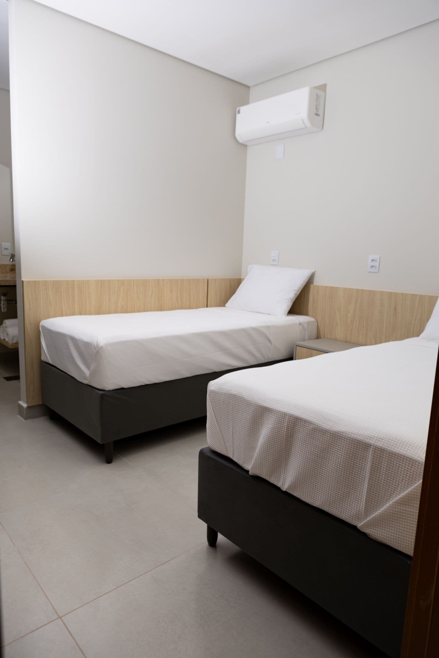 Habitación de hotel con dos camas, cabecero de madera, pared blanca y aire acondicionado instalado en el techo.