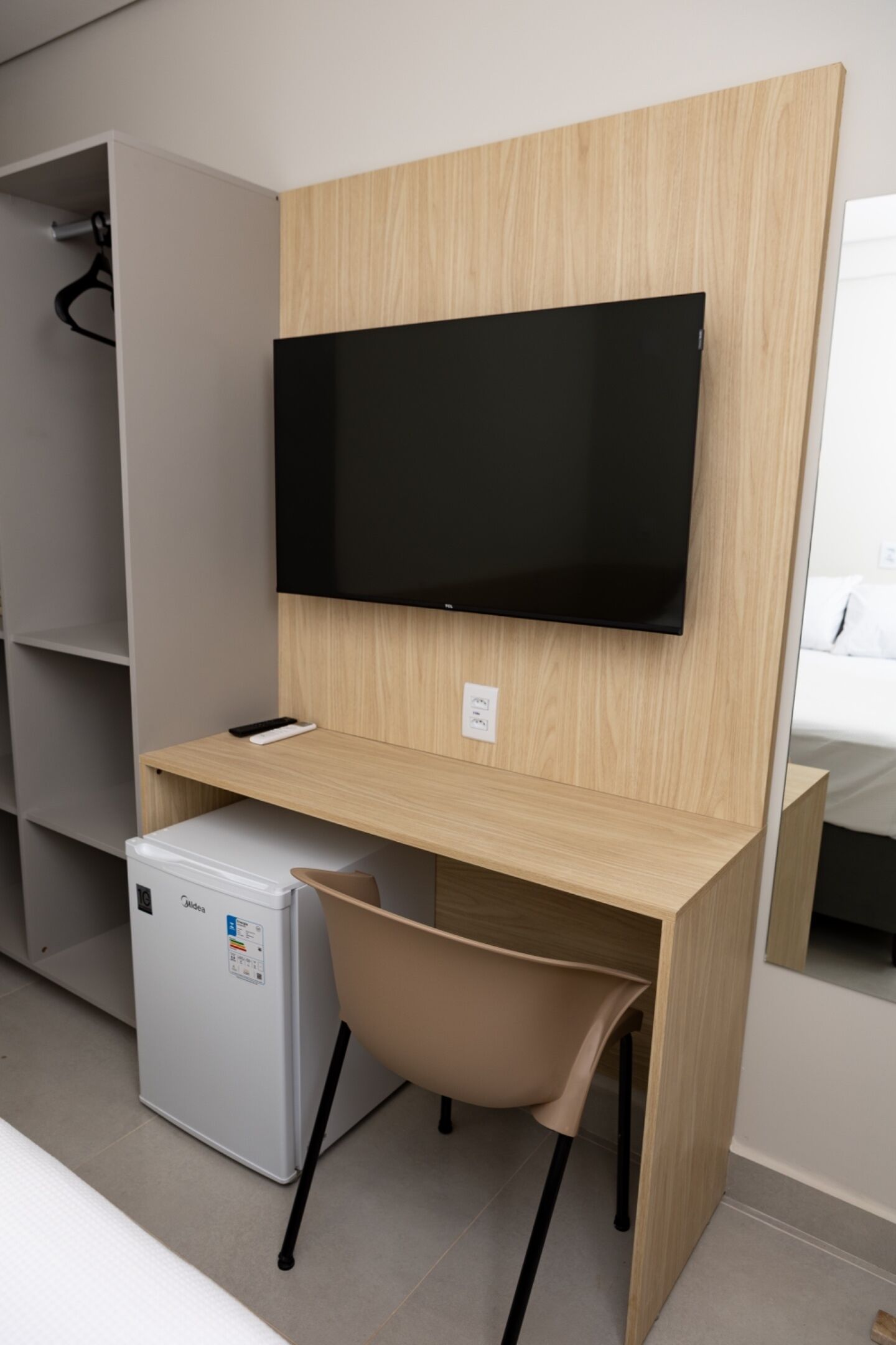 A compact room with a built-in desk, a television mounted above it, and a compact refrigerator to the left.