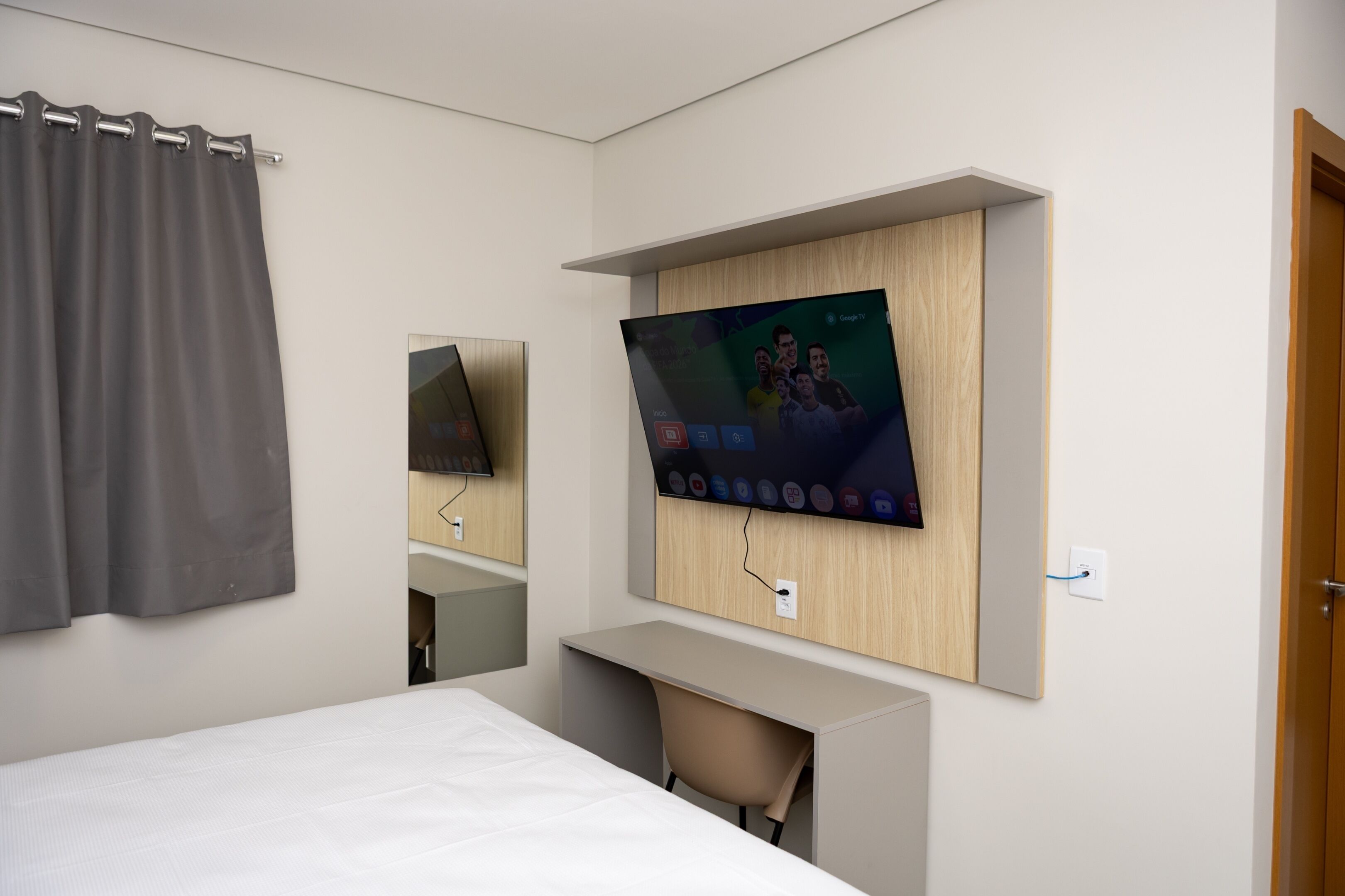 Ett hotellrum med en säng, ett skrivbord, en tv som är monterad på väggen och en gardin.