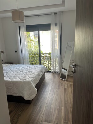 Ein Schlafzimmer mit einem Bett, Holzboden, einem Fenster mit Vorhängen und einer Tür mit Spiegel.