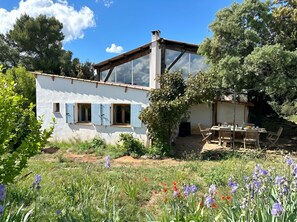 A white building with blue shutters, a patio with a table and chairs, and a garden with flowers. - Guest house immersed in nature with horses and donkeys (Jouques)