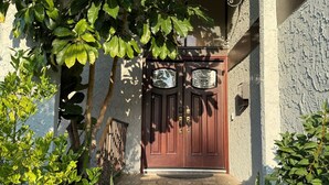 Une porte à deux battants en bois, ornée de deux plaques, un mur de pierre et un petit jardin avec un feuillage verdoyant.