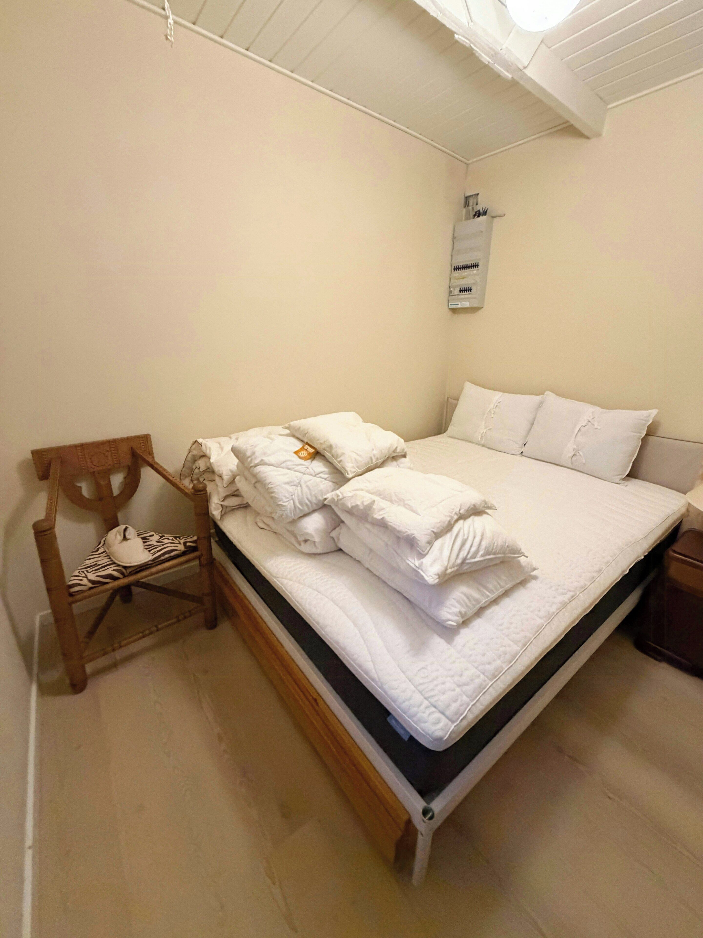 Uma cama bem arrumada com almofadas e um cobertor dobrado, uma cadeira de madeira com uma almofada estampada em zebra, e uma pequena mesa de cabeceira com um abajur.