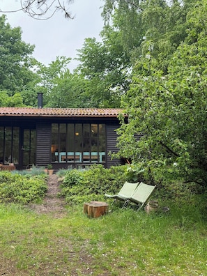 Ein modernes Haus mit großem Fenster, einer Holzterrasse und einem Gartenweg, der zu einer Bank führt.