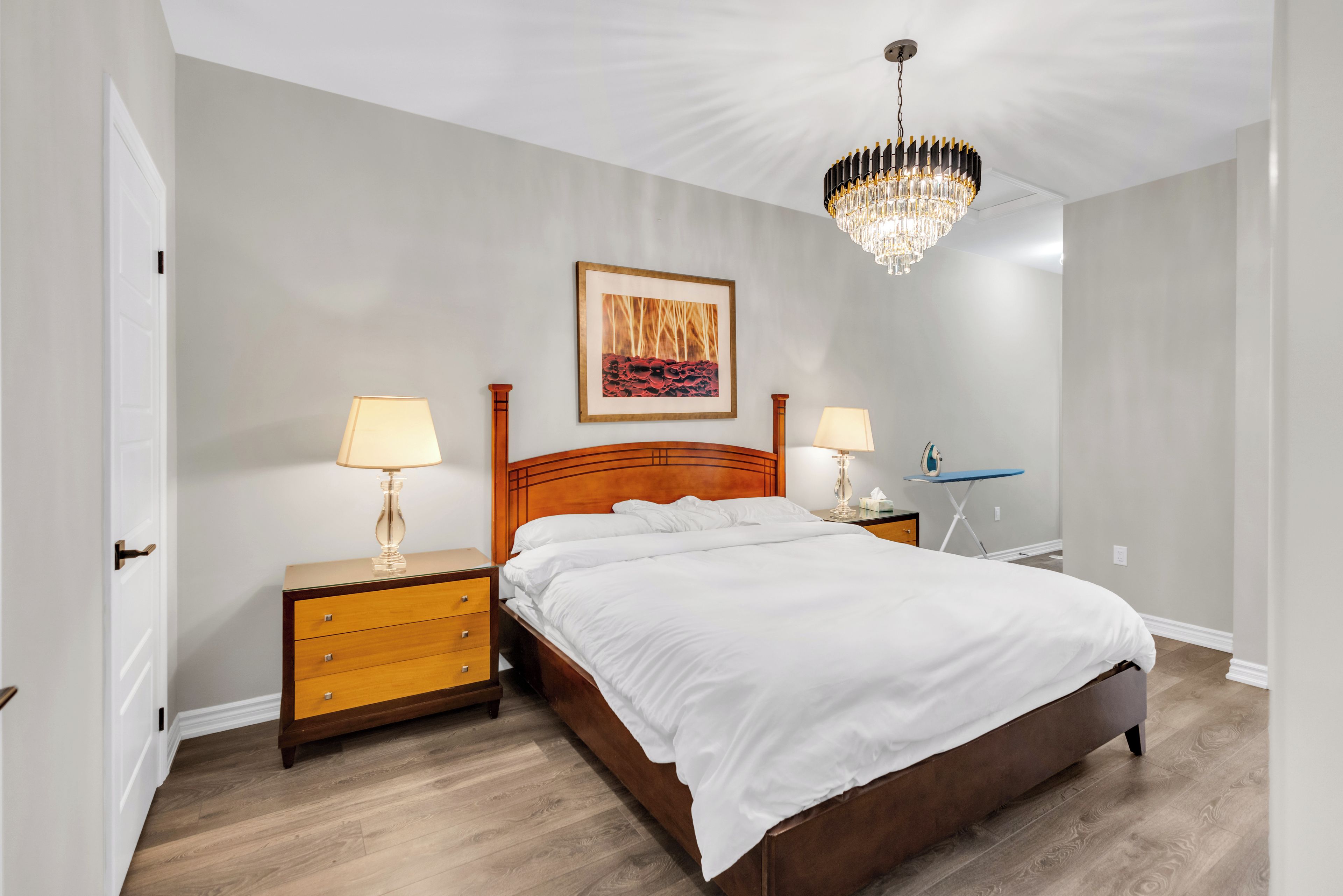 A hotel room with a wooden bed, two bedside tables with lamps, a chandelier, and a framed picture on the wall.