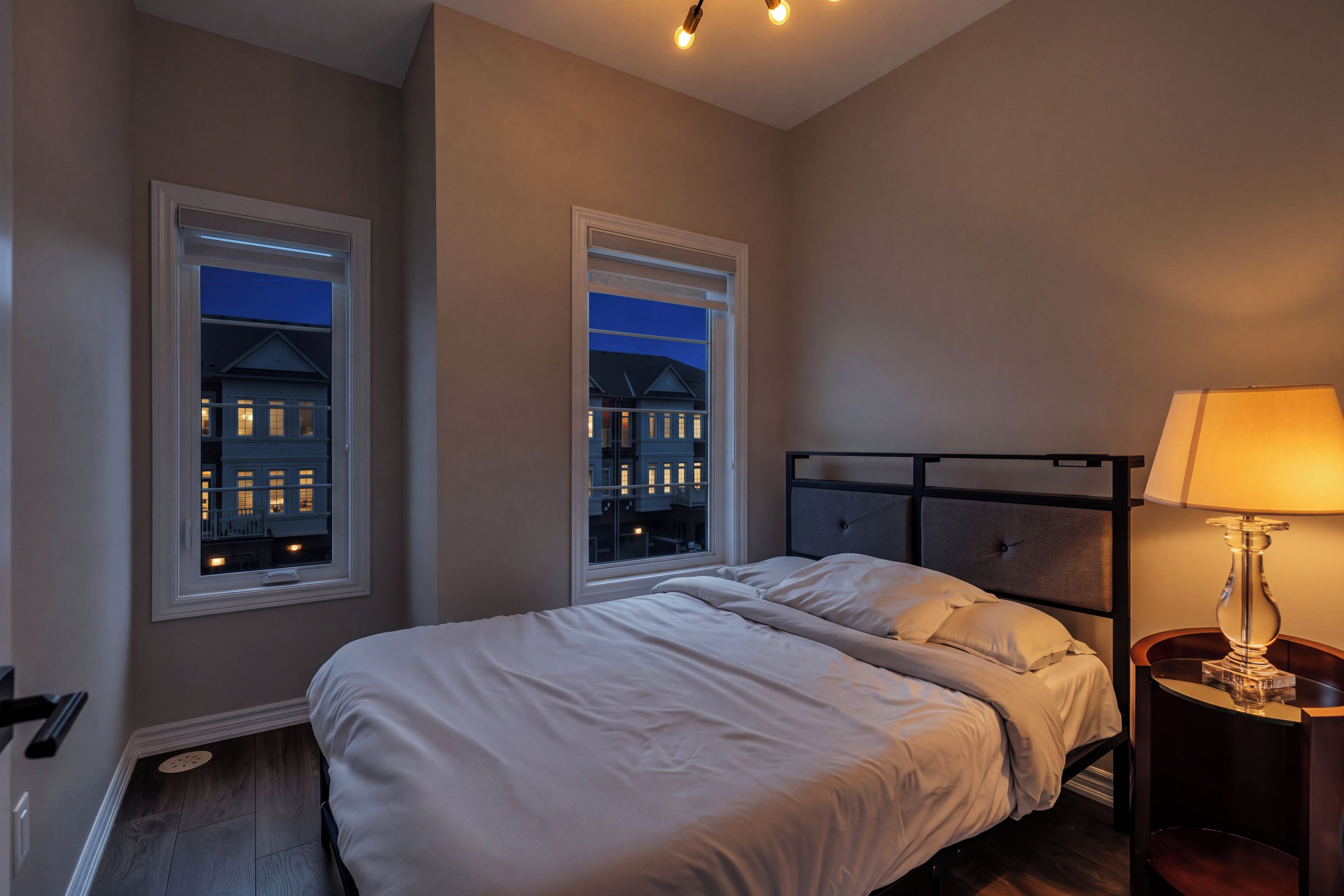 A bedroom with a bed, two windows, a nightstand, and a lamp.
