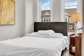 A neatly made bed with white linens in a room with large windows, a wall-mounted artwork, and a bedside table with a lamp. - Bright 3BR Home with Balcony & Free Parking (Bradford West Gwillimbury)