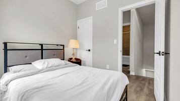 Habitación de hotel con cama, mesita de noche, lámpara, puerta y vista al baño.