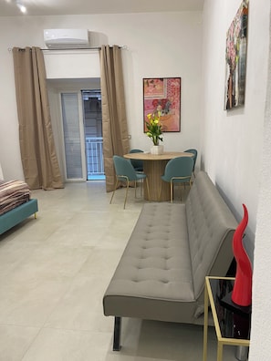 Ein Wohnzimmer mit einer Couch, einem Esstisch mit Stühlen, einer roten Vase und einem Bild an der Wand.