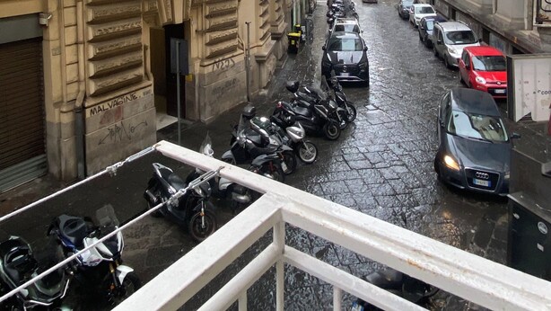 A narrow street with parked cars and motorcycles, buildings with balconies, and a wet pavement. - Basile House: elegance and comfort in the center of Naples (Napoli)