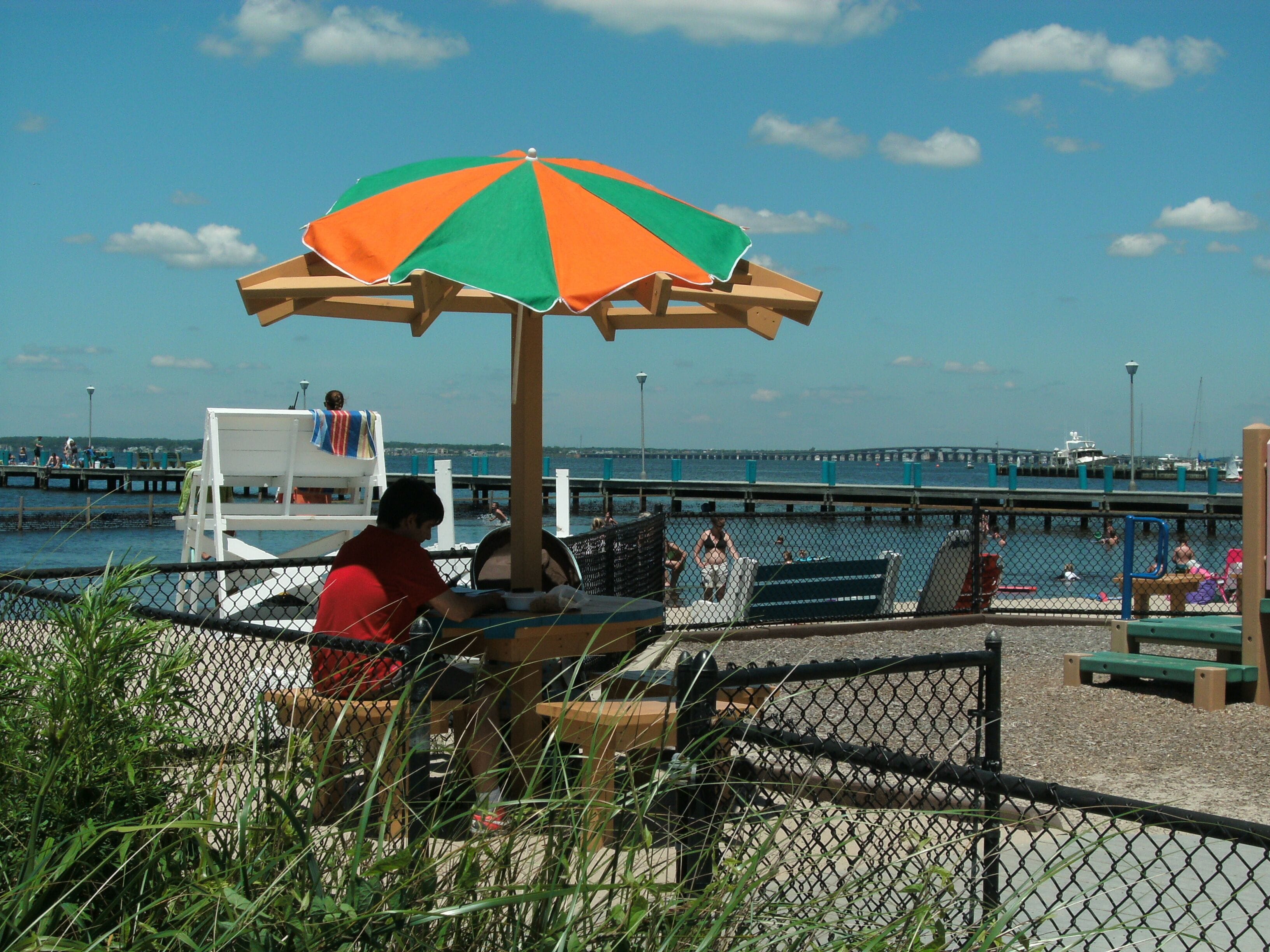 A beachside seating area with a wooden bench, a chain-link fence, and a view of the water and pier in the background.