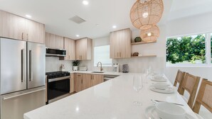 Une cuisine moderne dotée d’appareils électroménagers en acier inoxydable, de meubles en bois, d’un plan de travail en marbre blanc et d’un coin repas avec une longue table dressée pour six personnes.