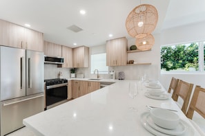 A modern kitchen with stainless steel appliances, wooden cabinets, a white marble countertop, and a dining area with a long table set for six. - Casa del Rio - 2BR on the Intracoastal (Boca Raton)