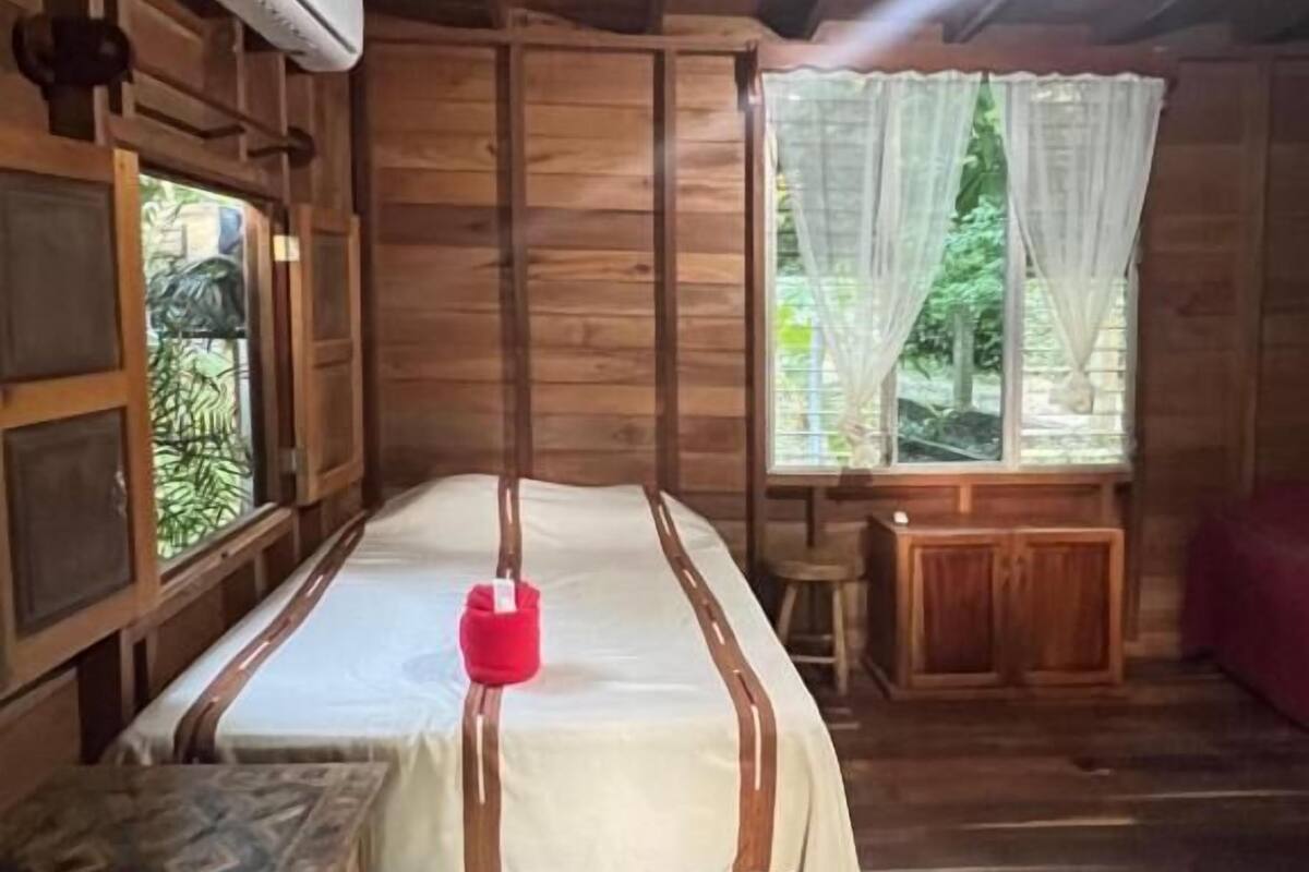 A wooden room with a bed, a bench, a window with curtains, and an air conditioning unit.