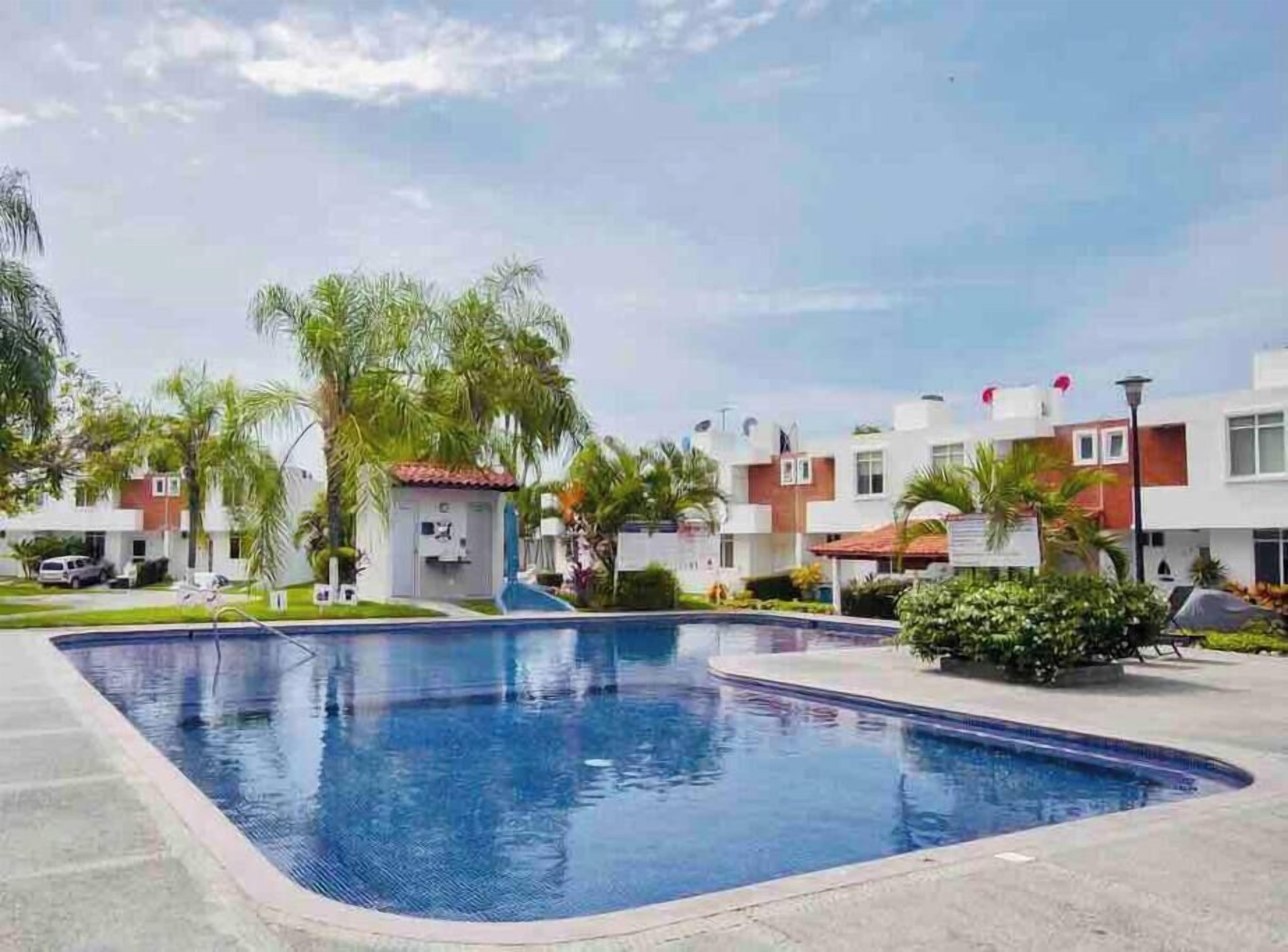 A swimming pool surrounded by modern buildings, palm trees, and a clear sky.