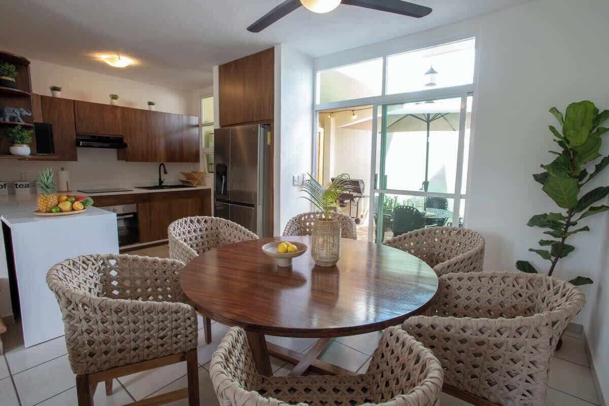 A modern kitchen with wooden cabinets, a stainless steel refrigerator, and a dining area with wicker chairs and a round wooden table.