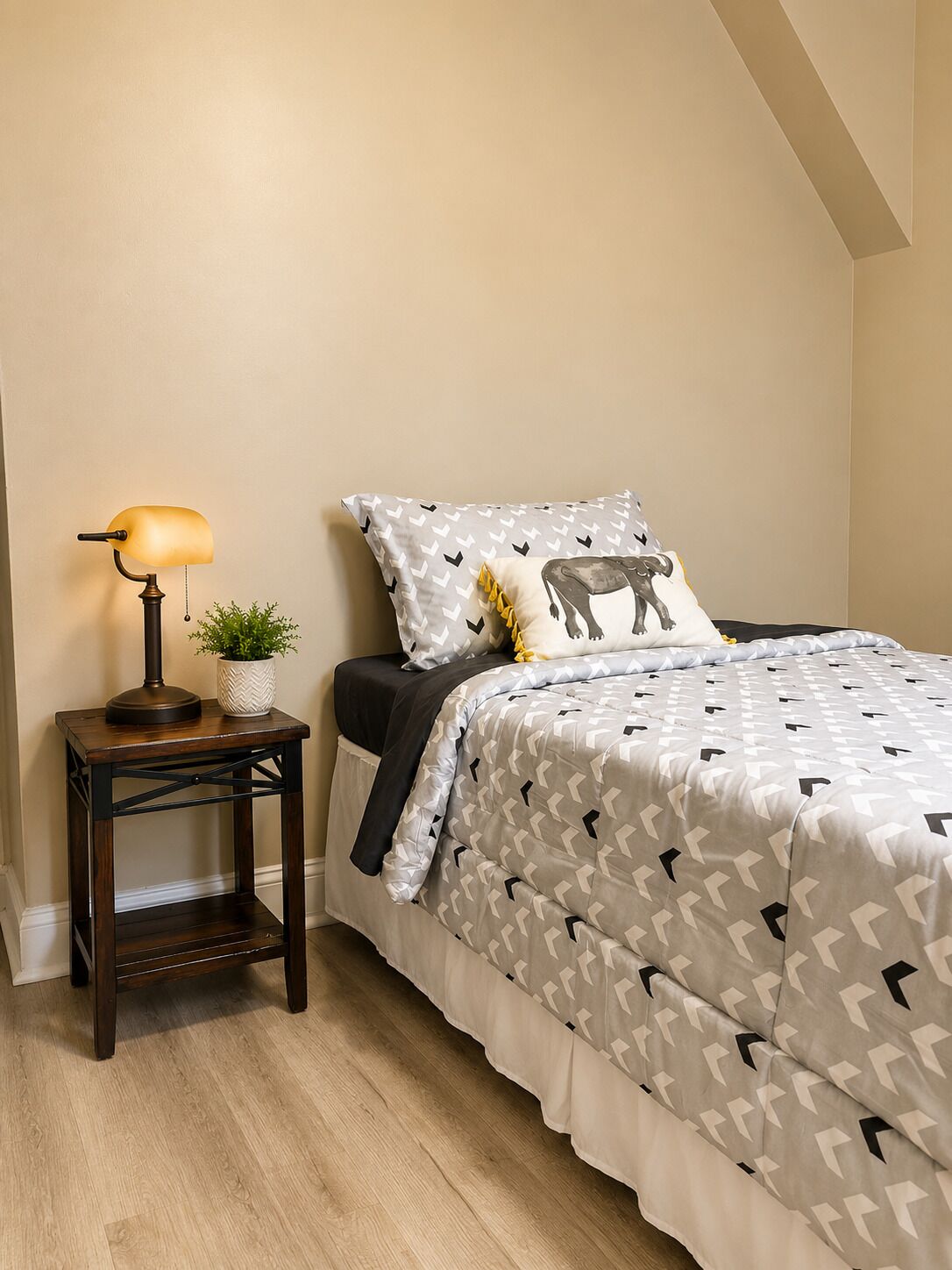 A bedroom with a bed, a bedside table with a lamp, a potted plant, and a patterned bedspread.