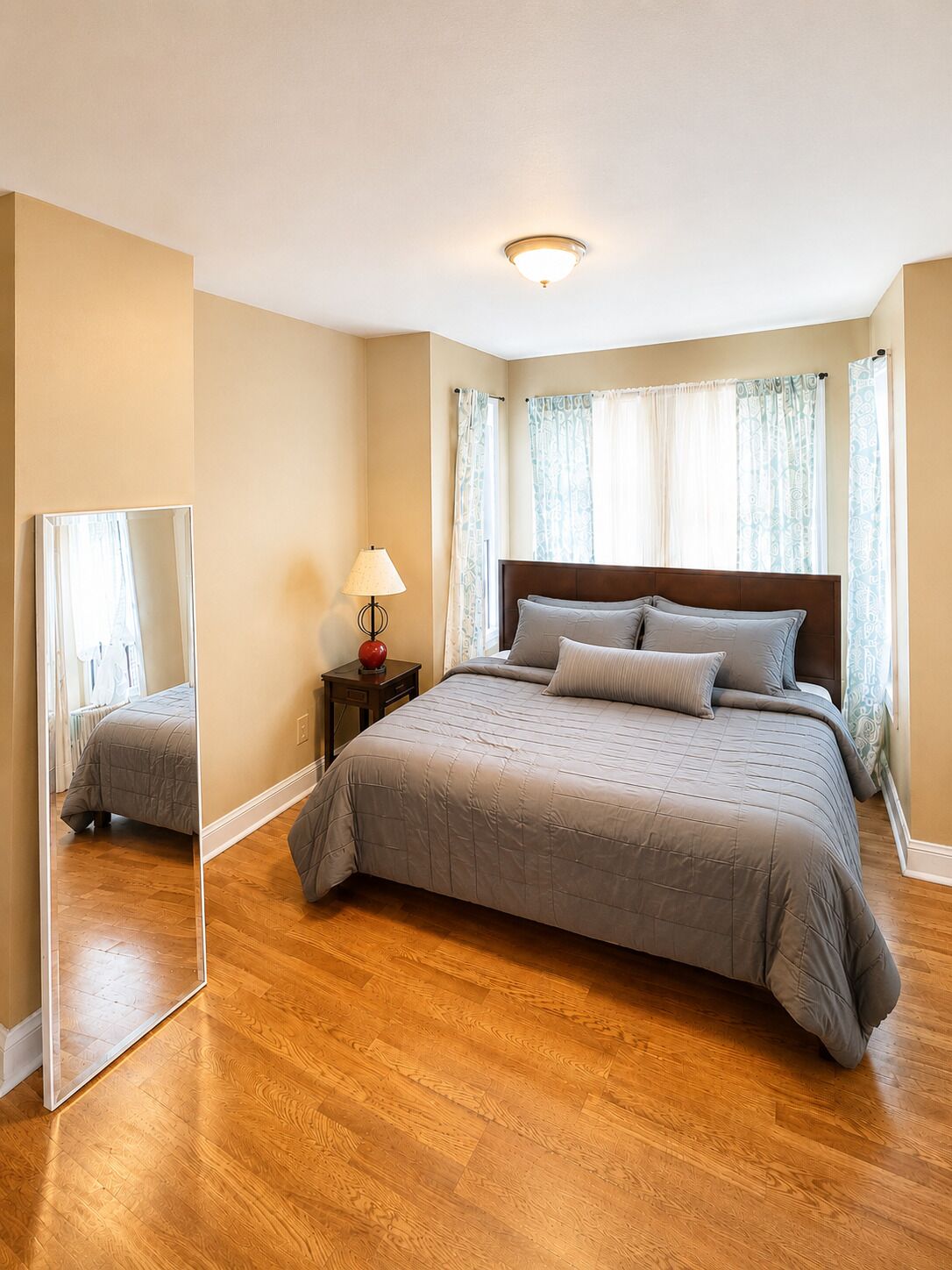 Un dormitorio con cama, mesita de noche, lámpara, espejo y ventana con cortinas.