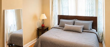 Un dormitorio con cama, mesita de noche, lámpara, espejo y ventana con cortinas.