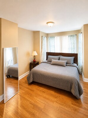 Ein Schlafzimmer mit einem Bett, einem Nachttisch, einer Lampe, einem Spiegel und einem Fenster mit Vorhängen.